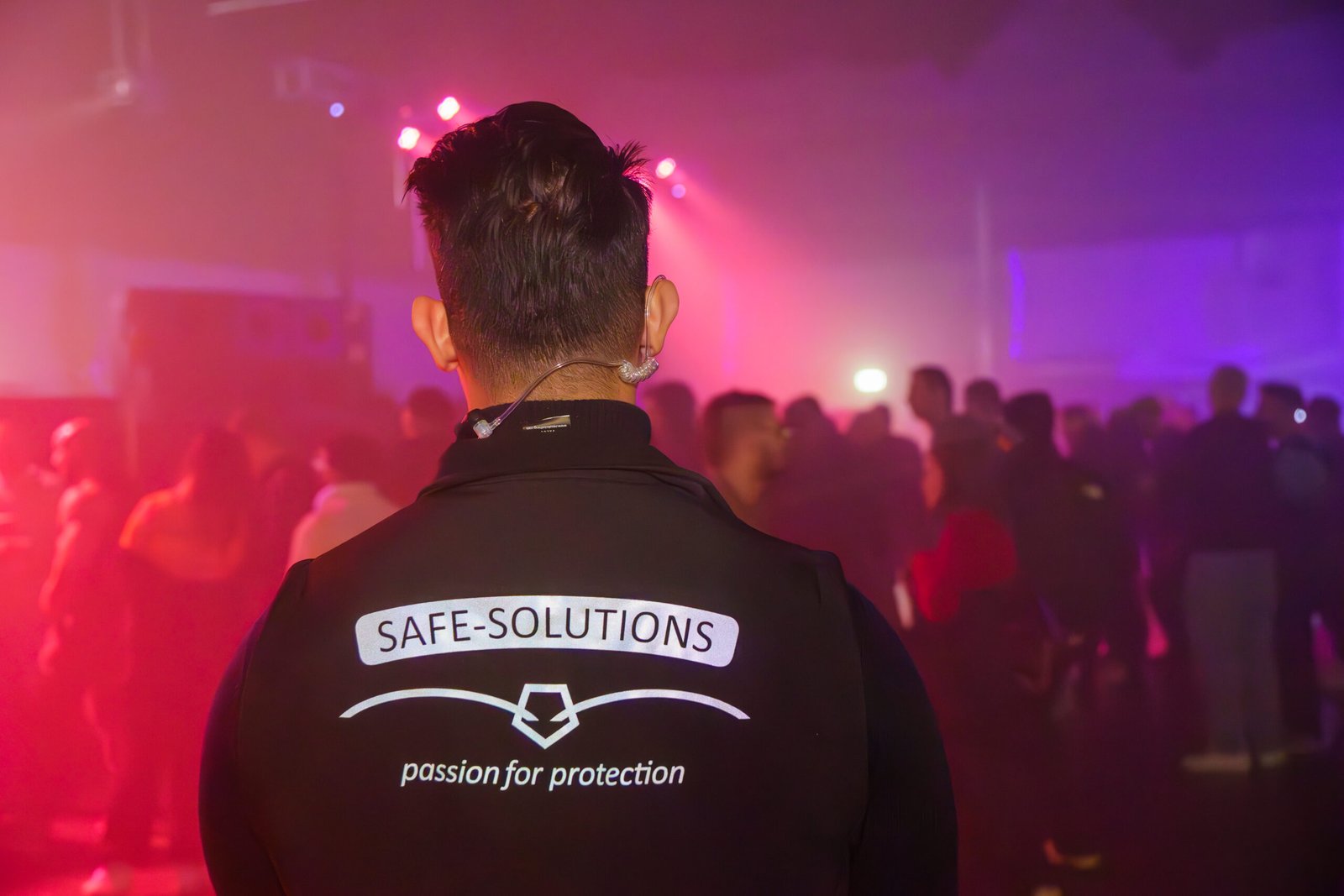 Safe Solutions guard seen from behind, watching a crowd under pink and purple lights